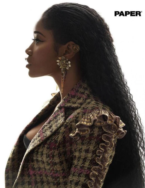Profile portrait of a Black woman with long curly hair, wearing a patterned tweed jacket, and large jeweled earrings against white.