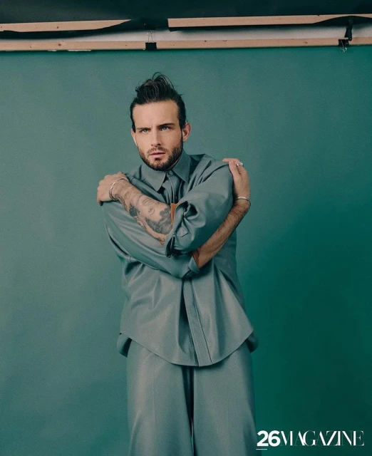 Portrait of a bearded man with tattooed arms wearing a gray shirt and trousers standing in front of a green studio backdrop.