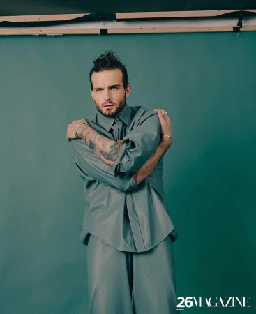 Portrait of a bearded man with tattooed arms wearing a gray shirt and trousers standing in front of a green studio backdrop.