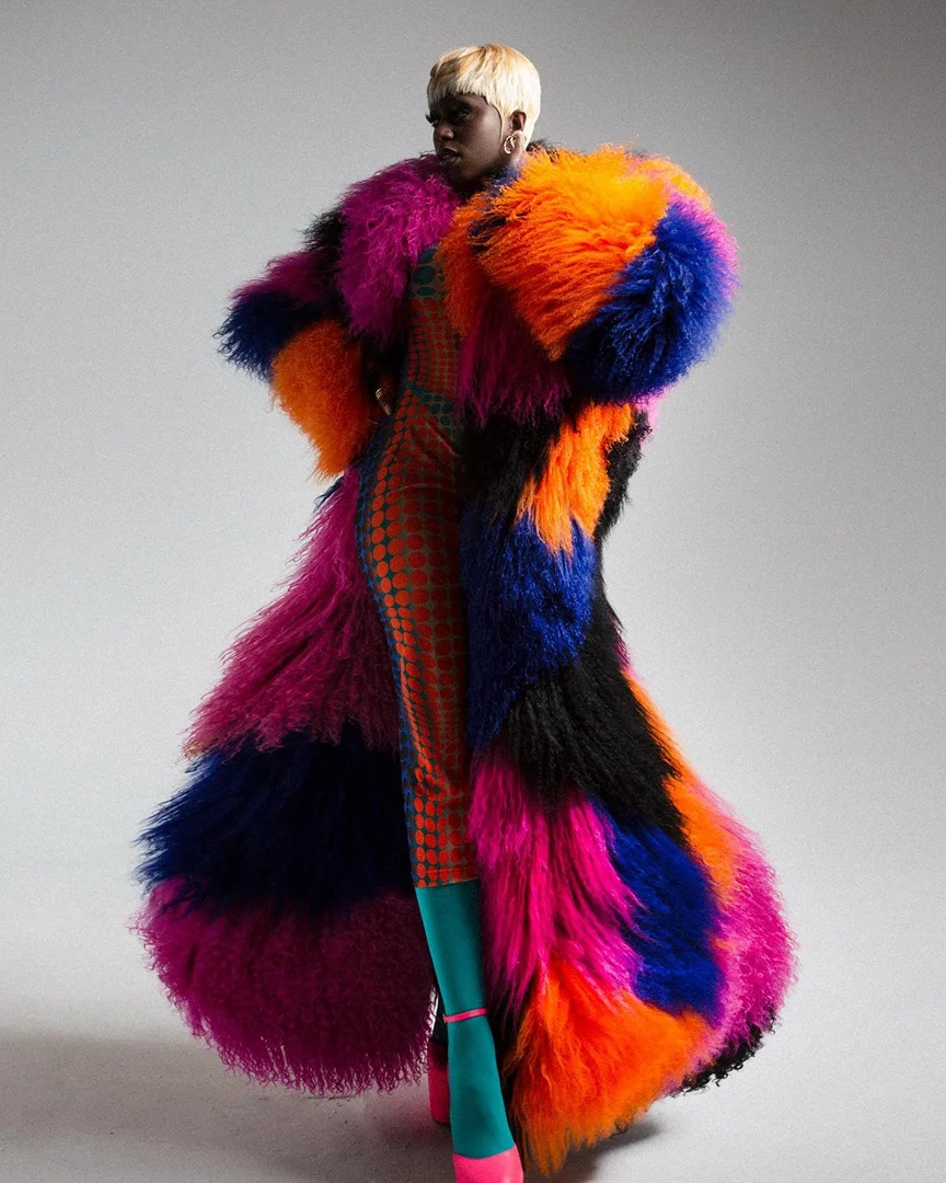 Woman poses in long, multicolored fur coat, patterned jumpsuit, teal socks, and pink heels against grey backdrop in full studio shot.