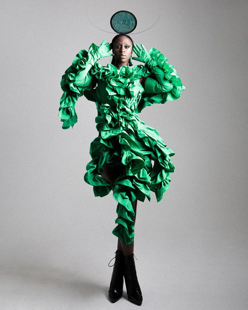 A dark-skinned adult female posing in a full shot, wearing a vivid green ruffled dress and black boots, against a light gray background.