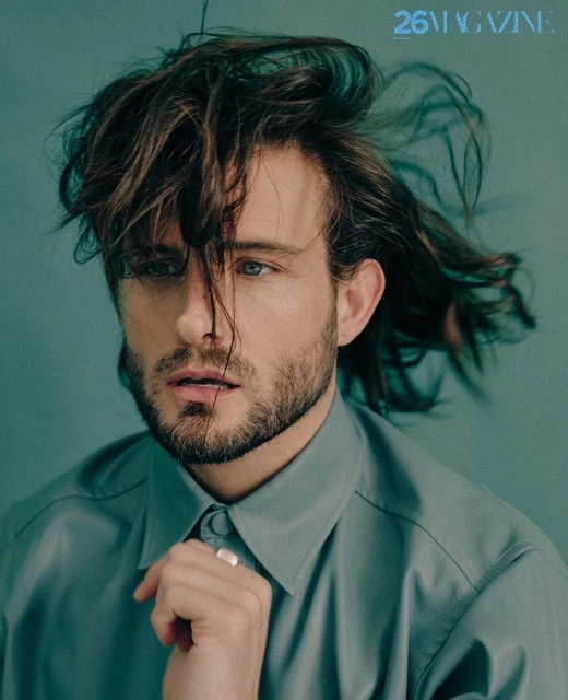 Portrait of a bearded, light-skinned male model with long, dark hair in a light blue-grey button-down shirt against a light blue background.
