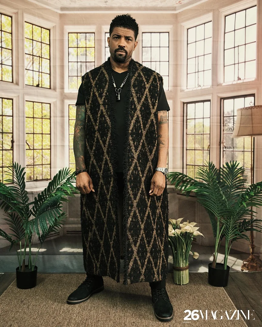 Full body portrait of an adult Black male in a patterned vest, standing indoors near a large window with potted plants.