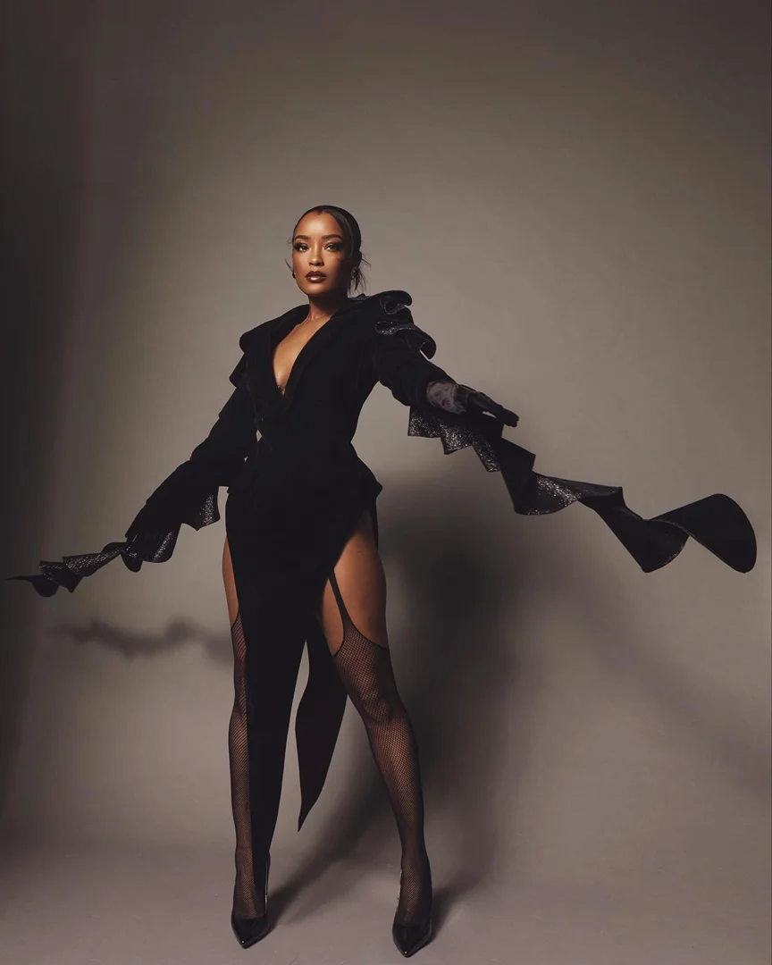Elegant woman in a black dress with flowing sleeves, fishnet stockings, and high heels, posing in a photo studio with a neutral background.