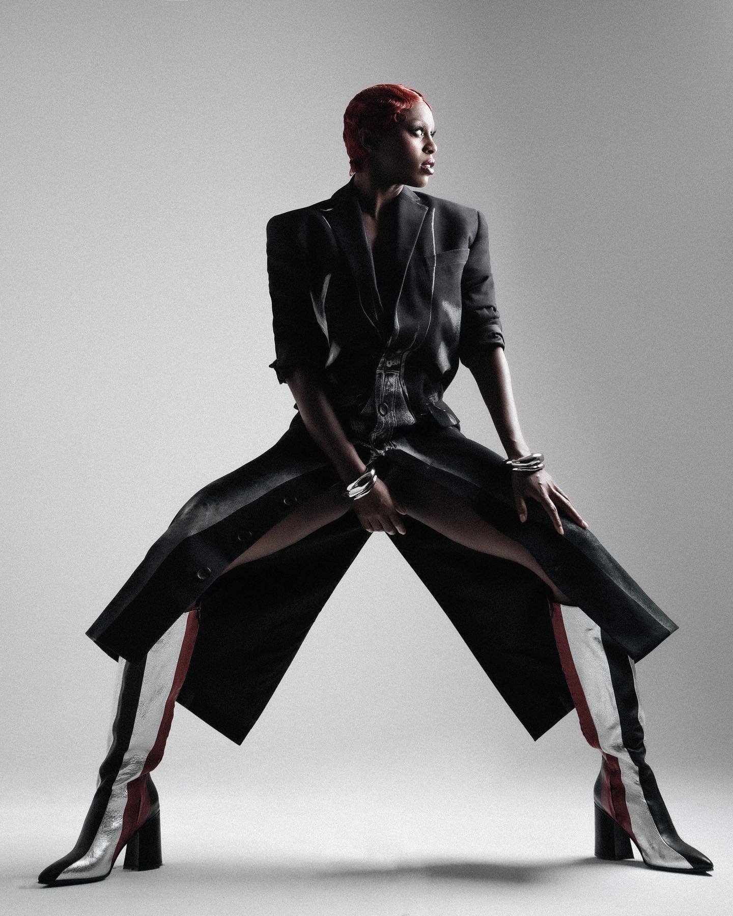Fashion shot of androgynous adult in an open black blazer jacket, sitting with legs spread, wearing silver, red, and black boots.