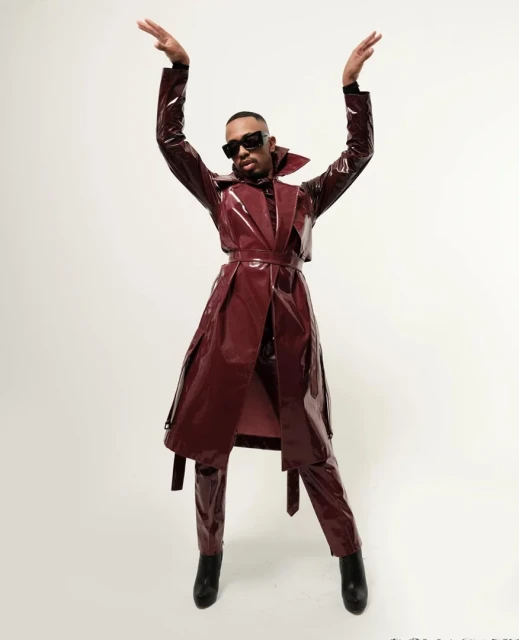 Adult male poses in a shiny burgundy coat and pants with arms raised against an off-white backdrop in a studio setting.