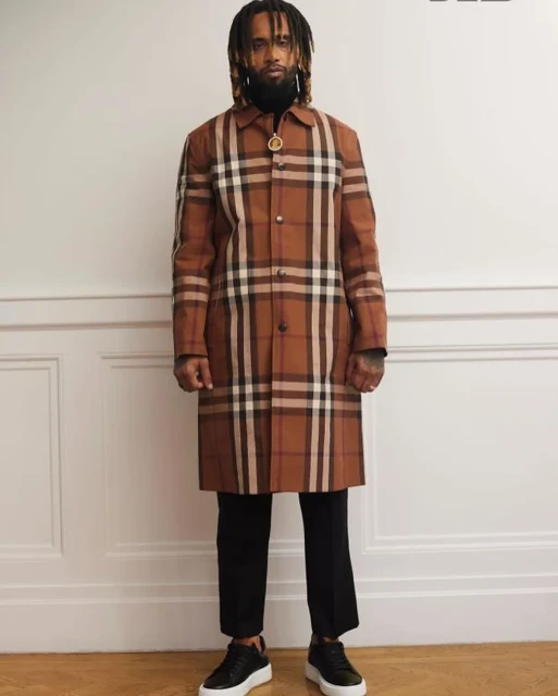 Man stands indoors wearing a Burberry plaid trench coat, black pants, and black leather sneakers against a white paneled wall.