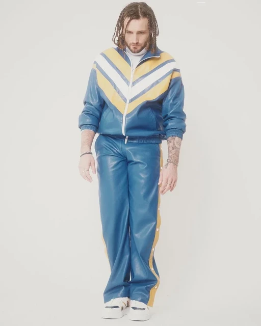 Full-length portrait of a man with dreadlocks in a blue and yellow tracksuit against a white background, posing for a fashion shot.