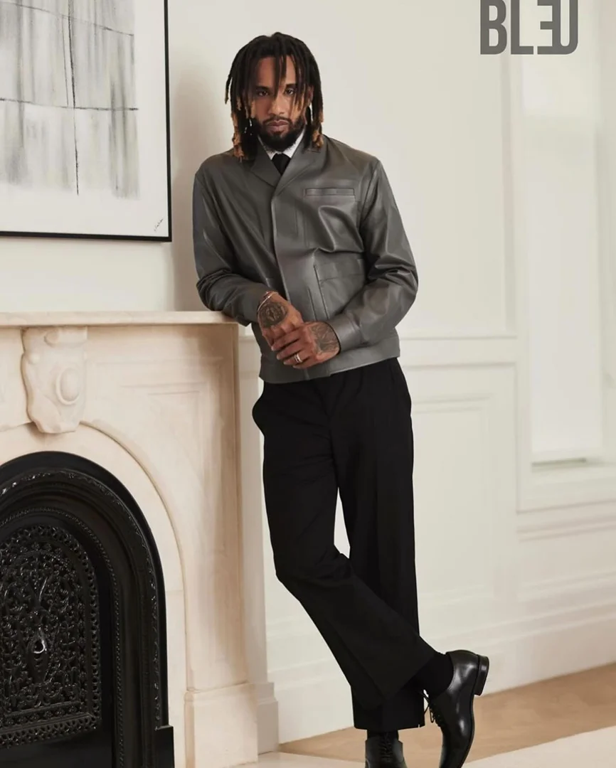 Stylish man poses indoors, wearing a gray leather jacket, black pants, and dress shoes, next to a fireplace, under soft, diffused light.