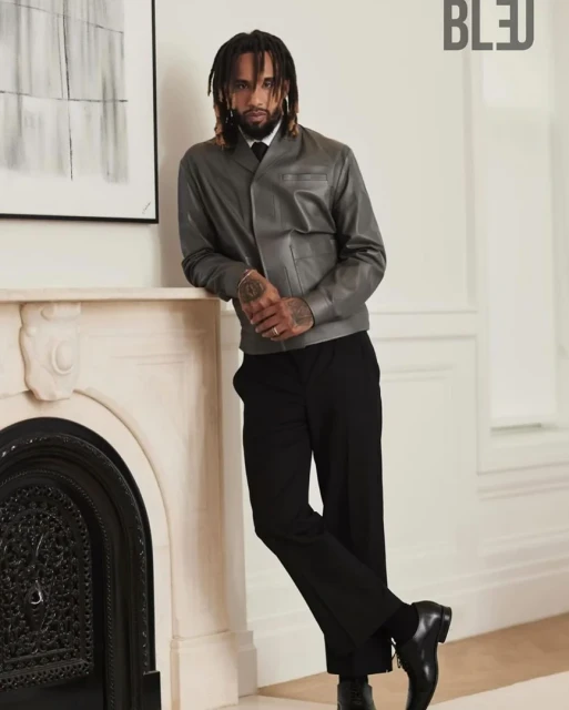 Stylish man poses indoors, wearing a gray leather jacket, black pants, and dress shoes, next to a fireplace, under soft, diffused light.