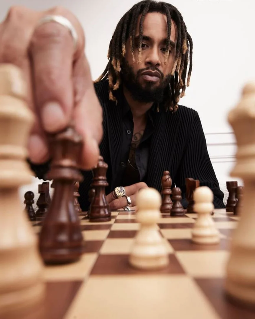 A man with dreadlocks playing chess, capturing a chess piece on a wooden chessboard in a brightly lit indoor environment.