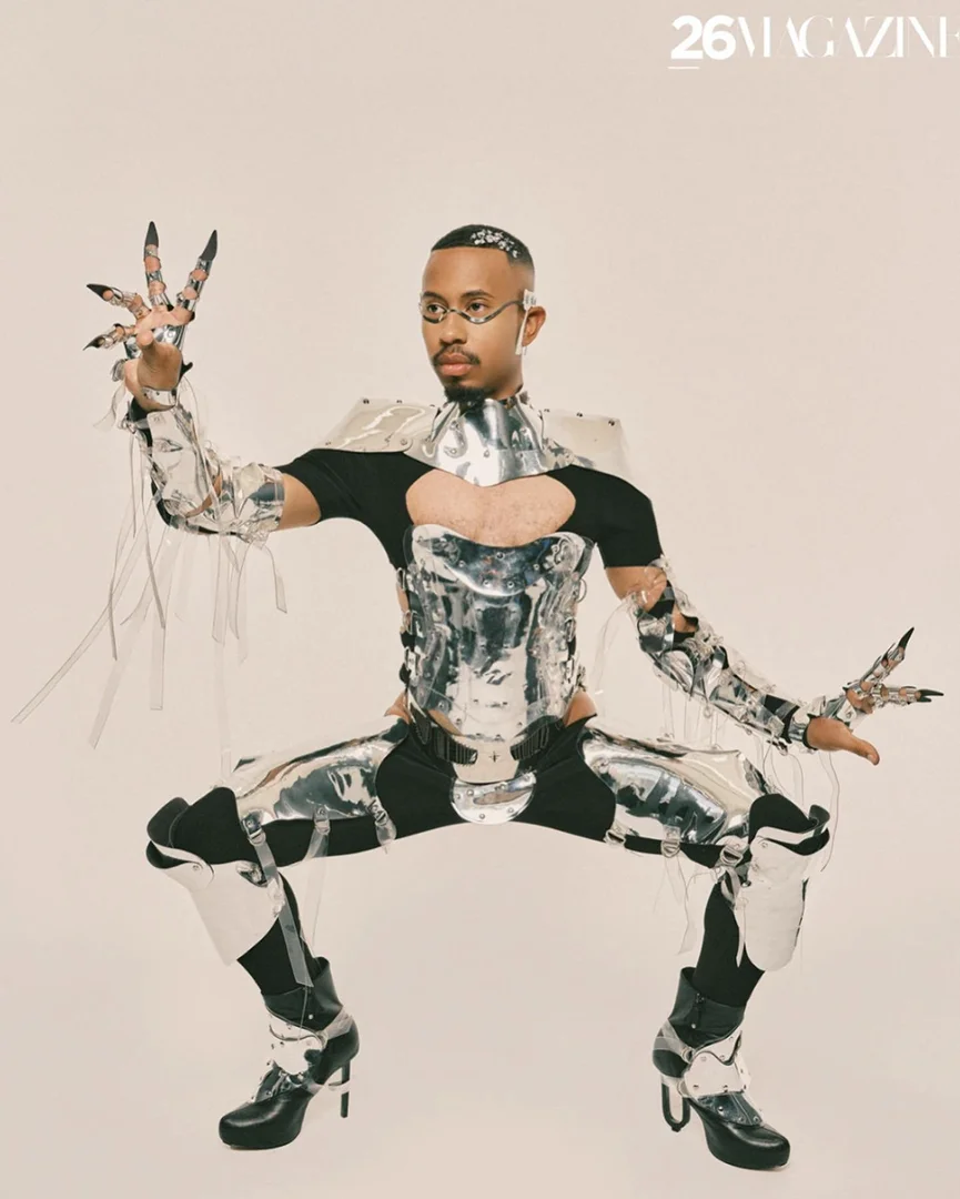 Adult male in reflective silver futuristic costume with glasses poses in a studio with unique hand jewelry and black high-heel boots.
