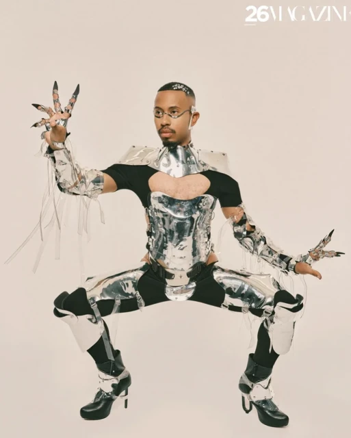 Adult male in reflective silver futuristic costume with glasses poses in a studio with unique hand jewelry and black high-heel boots.