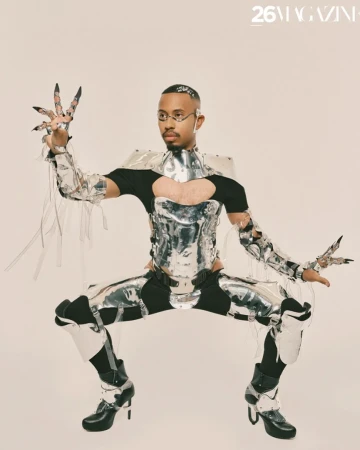 Adult male in reflective silver futuristic costume with glasses poses in a studio with unique hand jewelry and black high-heel boots.