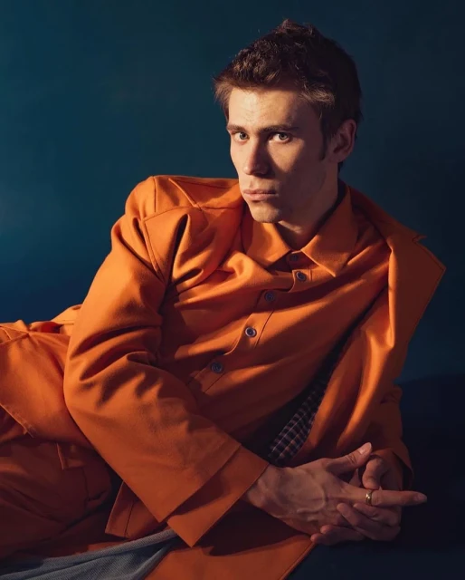 Portrait of an adult male wearing an orange shirt and pants, lying down against a dark blue backdrop in a photo studio setting.
