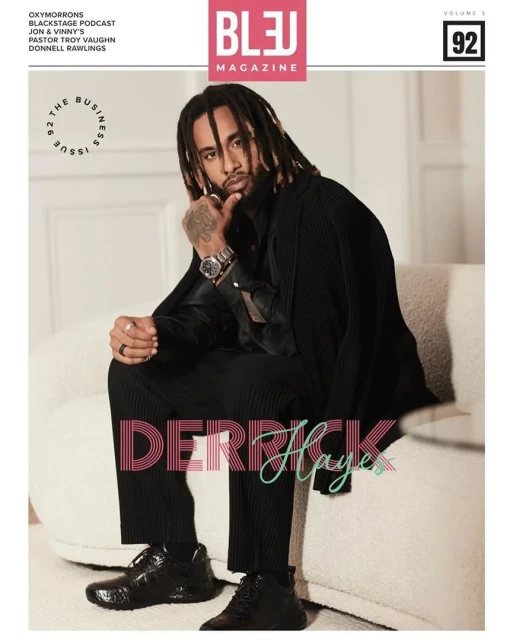 Magazine cover featuring a Black male model with dreadlocks wearing a black suit, sitting on a cream-colored chair, studio lighting.