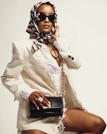 A woman in a white blazer and striped headscarf poses with a black clutch and gold jewelry against a light background in a fashion shot.
