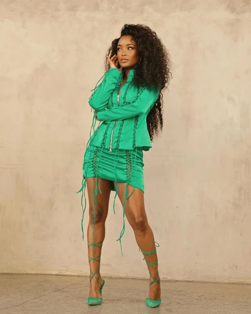 A confident Black woman poses in a green skirt suit and matching heels against a neutral backdrop, showcasing fashion and style.