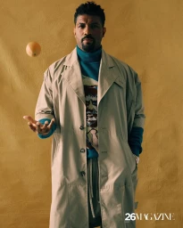 Man wearing a long beige trench coat over a blue turtleneck and tossing a small fruit in the air against a textured gold background