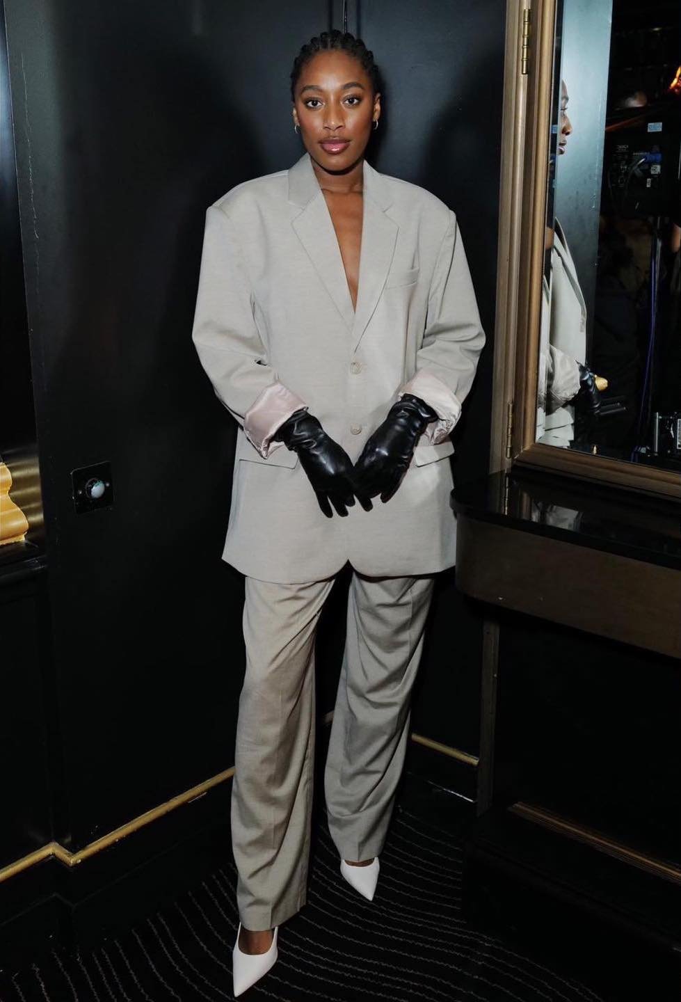 Stylish Black woman in an oversized grey suit, black gloves and white high heels in a dark interior setting, fashion portrait.