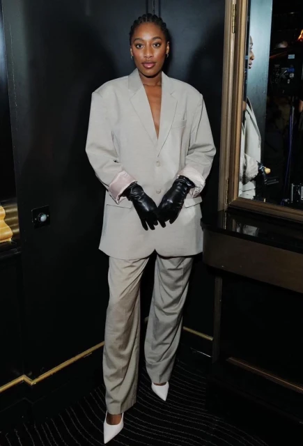 Stylish Black woman in an oversized grey suit, black gloves and white high heels in a dark interior setting, fashion portrait.