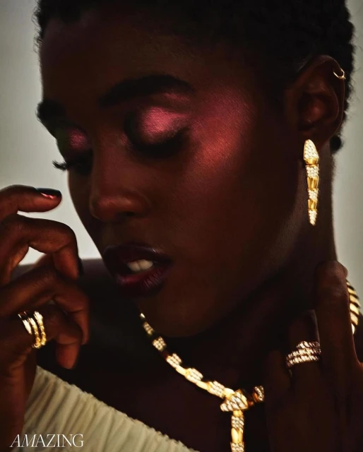 Close-up of a dark-skinned young woman with magenta eyeshadow wearing a gold, snake-themed jewelry set consisting of necklace, earrings, rings.