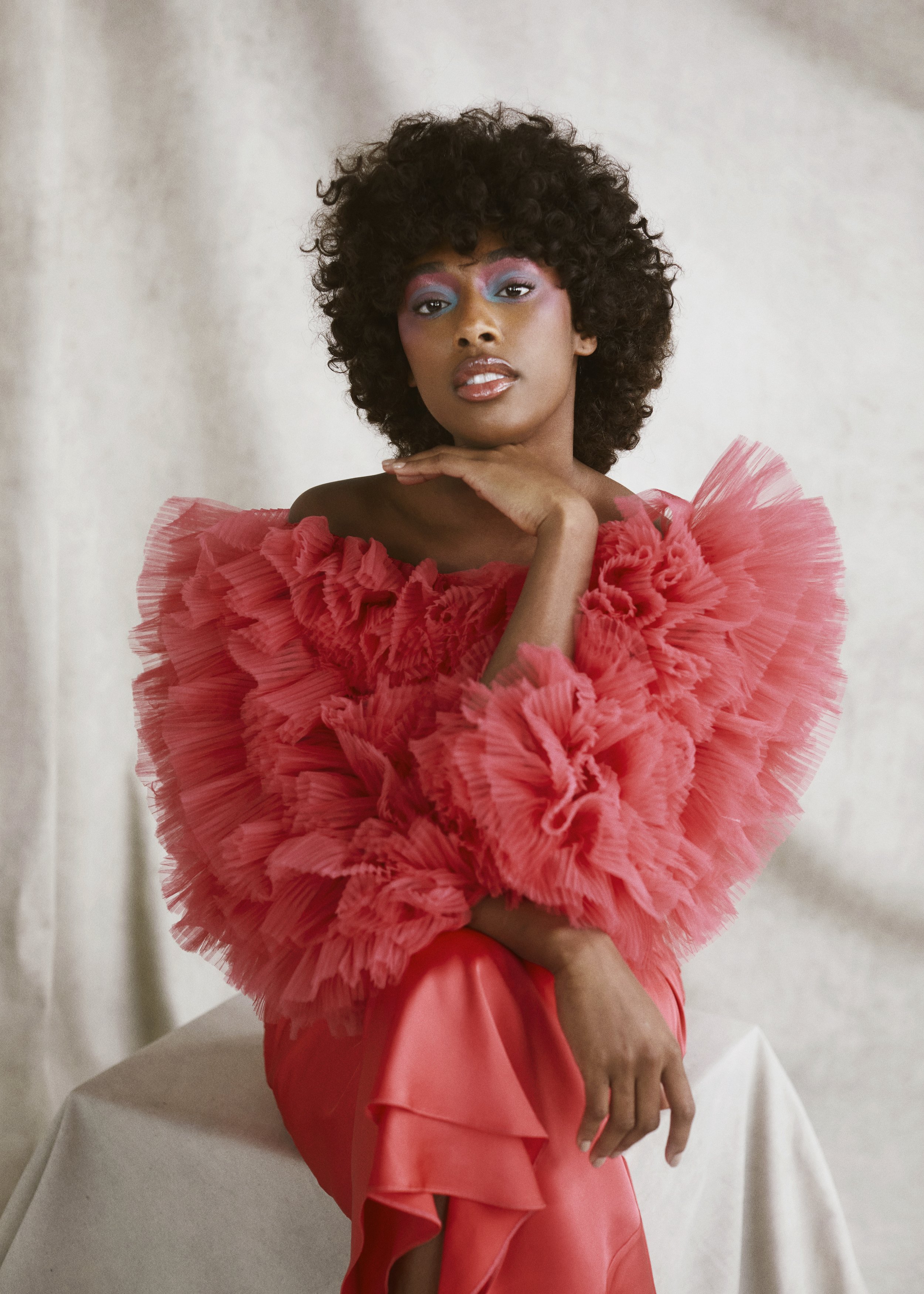 Portrait of an African American woman wearing a vibrant pink ruffled top and makeup, set against a soft, neutral backdrop.