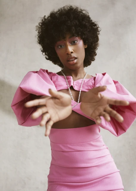 Portrait of a Black woman in a pink cut-out dress, accessorized with a beaded necklace, posing with outstretched hands in a studio setting.