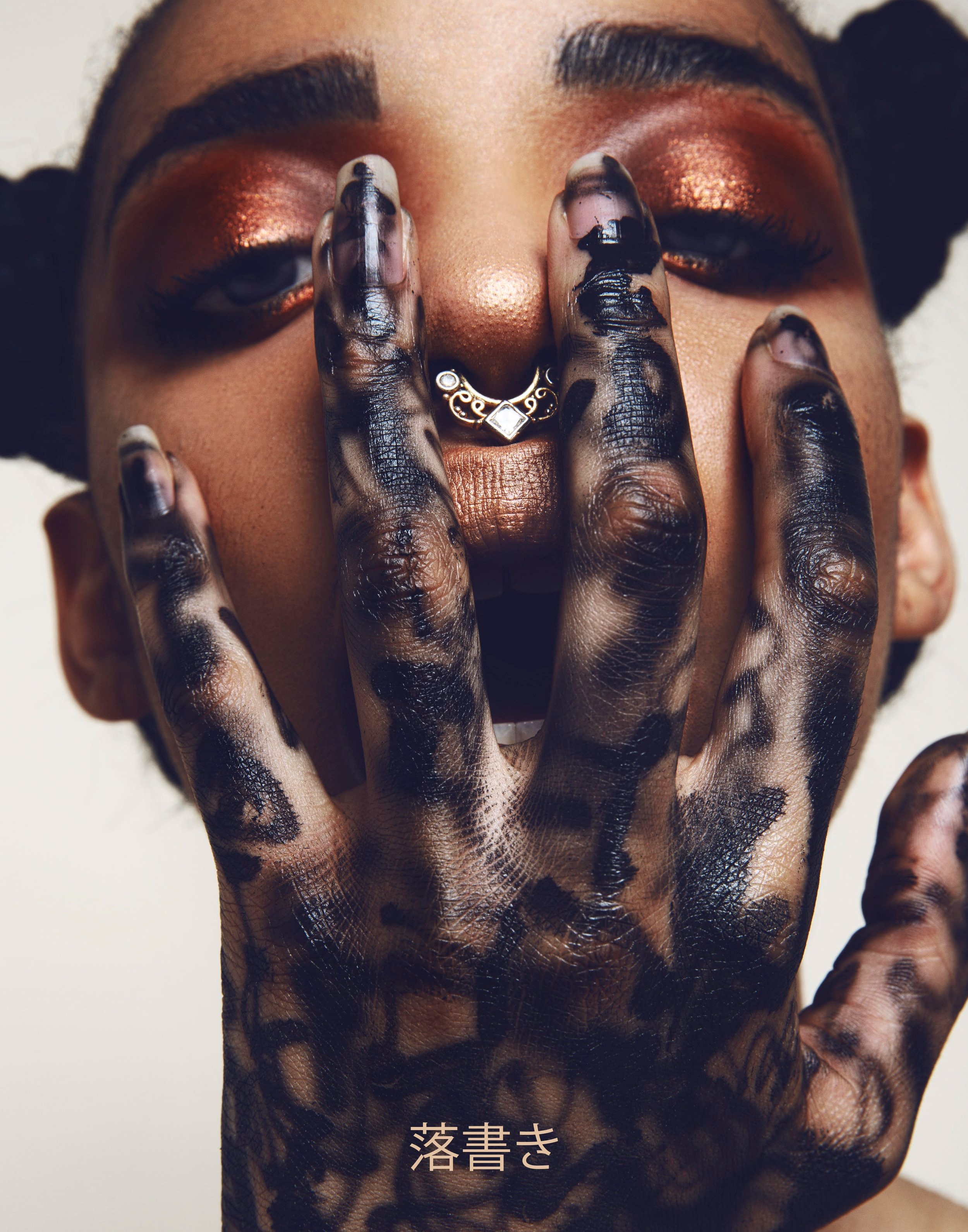 Eye-level portrait of a young adult woman with bronze makeup and styled hair, her hand featuring a black pattern is near her face.