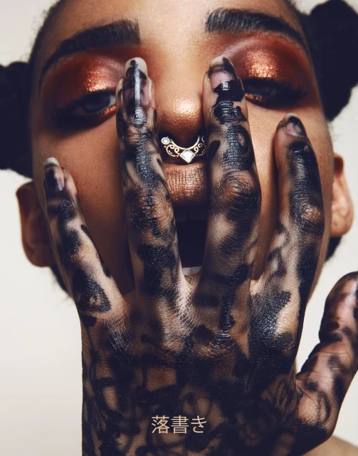 Eye-level portrait of a young adult woman with bronze makeup and styled hair, her hand featuring a black pattern is near her face.
