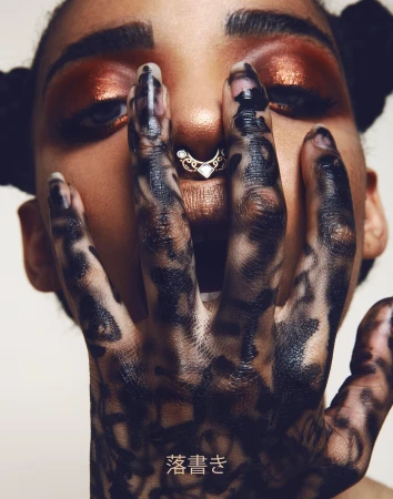 Eye-level portrait of a young adult woman with bronze makeup and styled hair, her hand featuring a black pattern is near her face.
