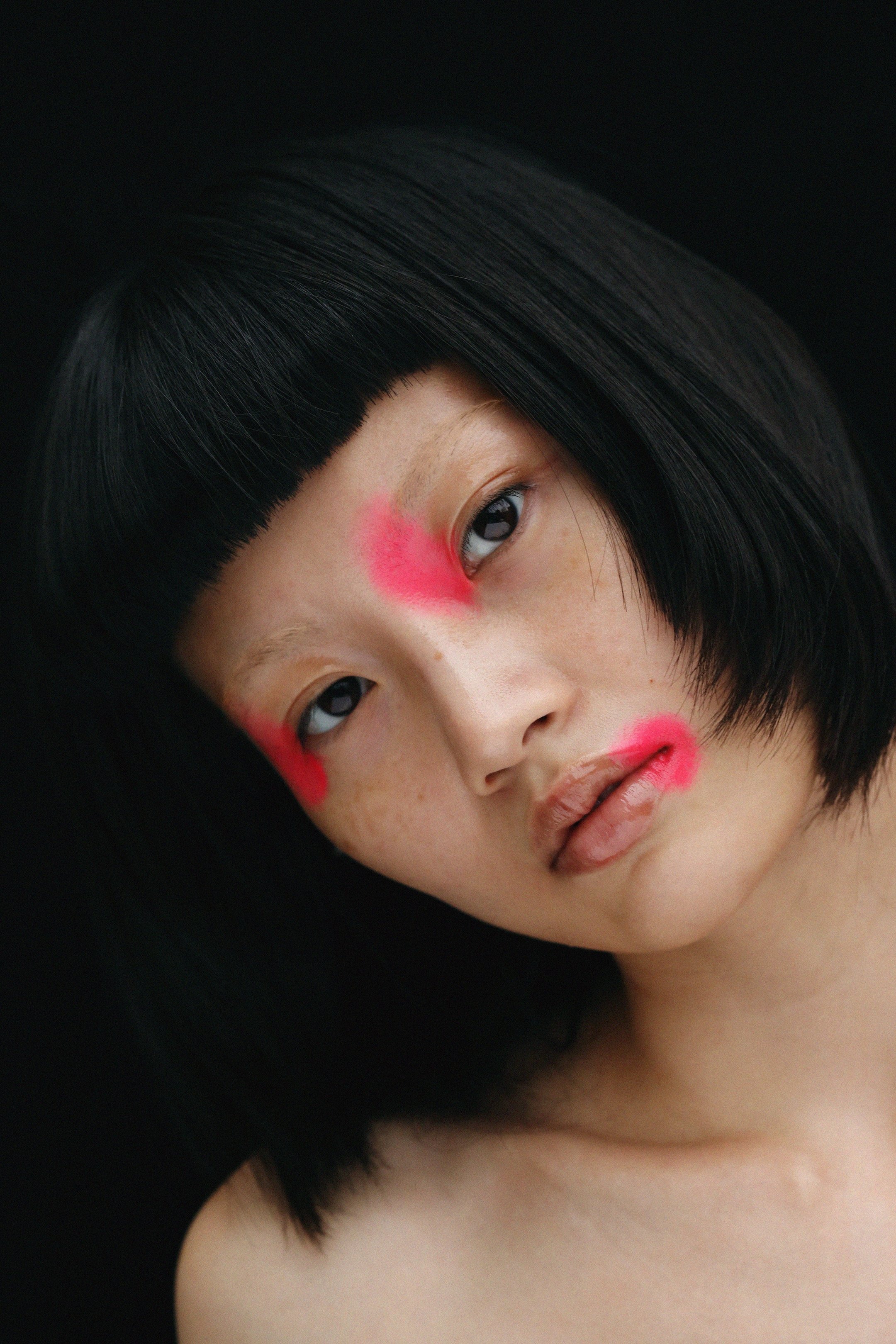 Close-up portrait of a woman with a dark bob haircut and striking neon pink makeup on her nose and lips against a black background.