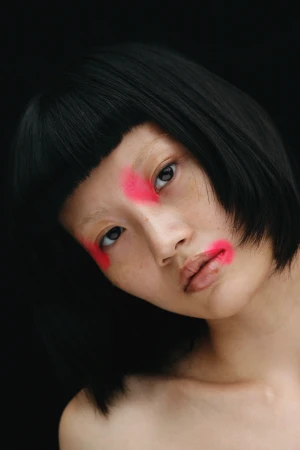 Close-up portrait of a woman with a dark bob haircut and striking neon pink makeup on her nose and lips against a black background.