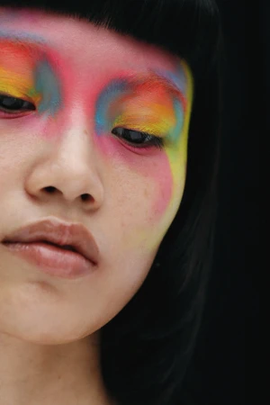 Close-up portrait of a woman wearing vibrant rainbow eyeshadow art and black blunt bangs against a dark background