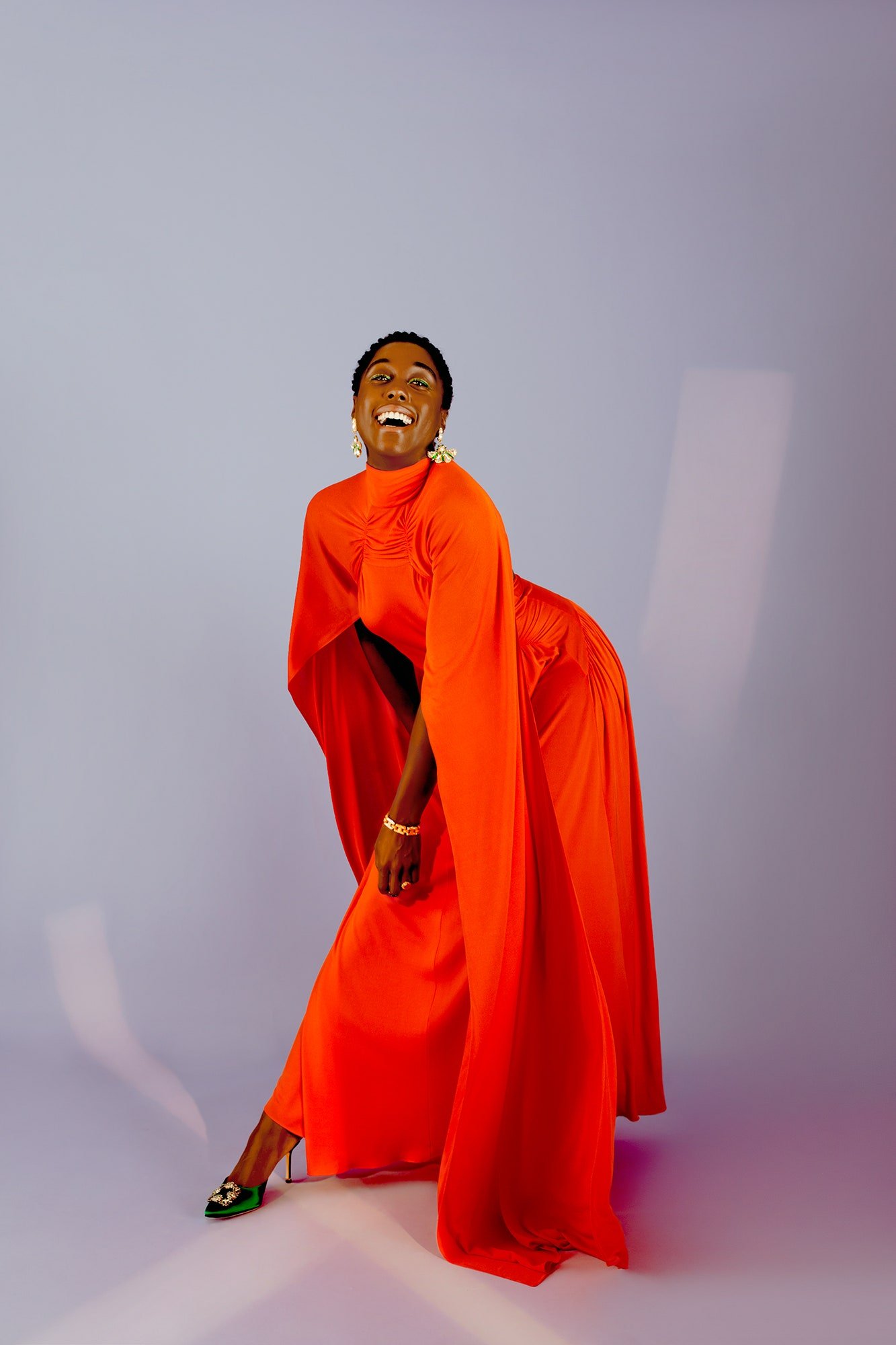 Full shot of an adult woman smiling and posing in a vibrant orange dress and green heels against a light purple backdrop in studio lighting.