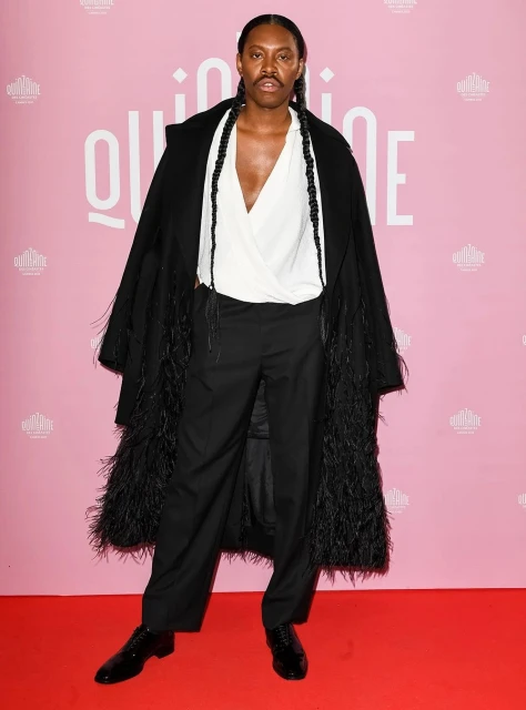 A person stands on a red carpet wearing a white blouse, black pants, a black coat with feathered trim, and black shoes.