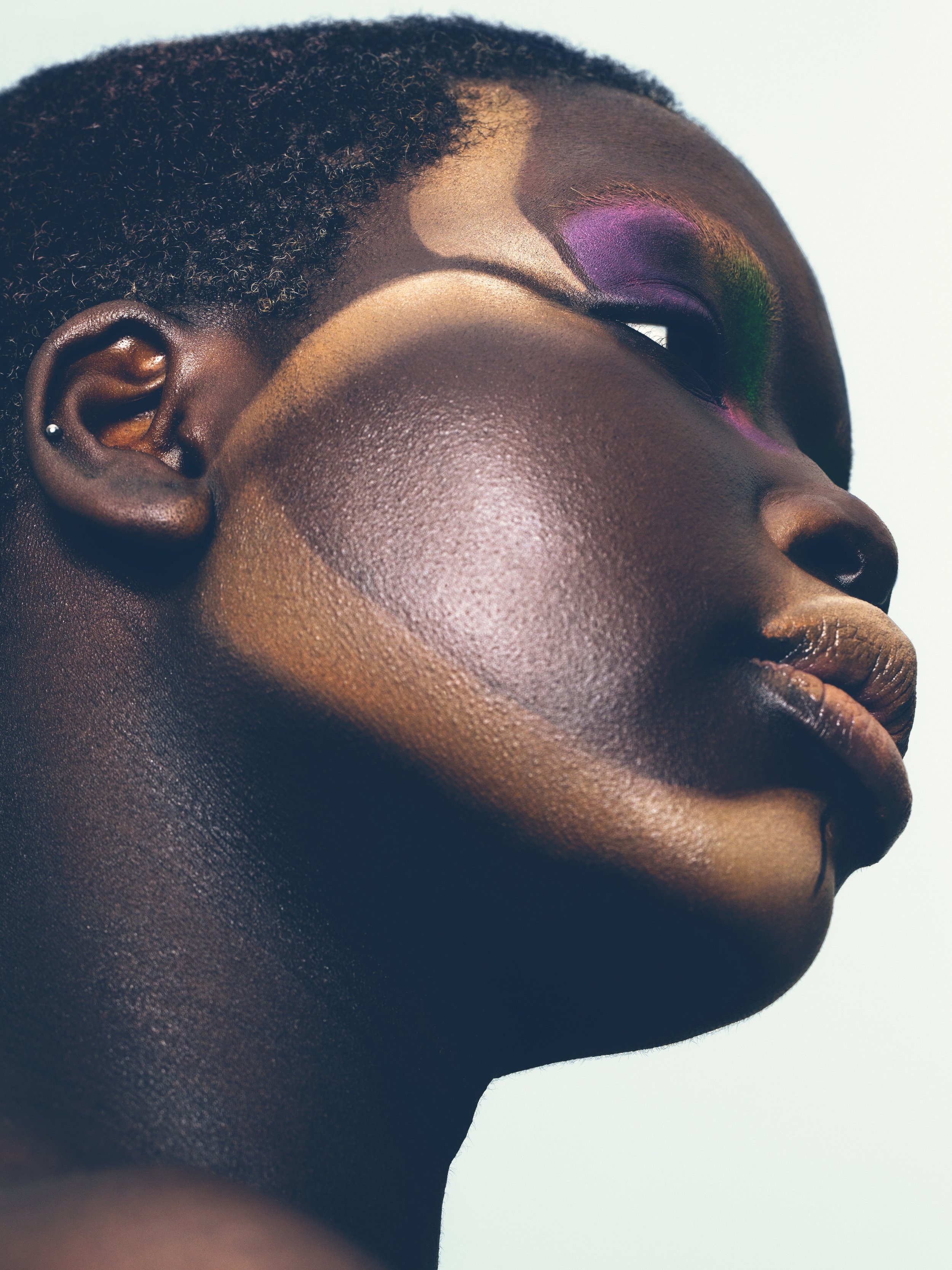 Close-up profile portrait of a Black woman with purple eyeshadow, dark skin, and short hair against a light background, showcasing beauty.