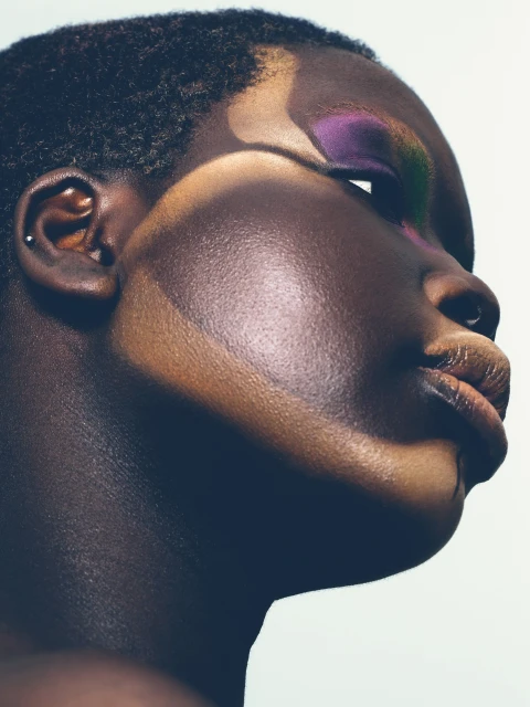 Close-up profile portrait of a Black woman with purple eyeshadow, dark skin, and short hair against a light background, showcasing beauty.