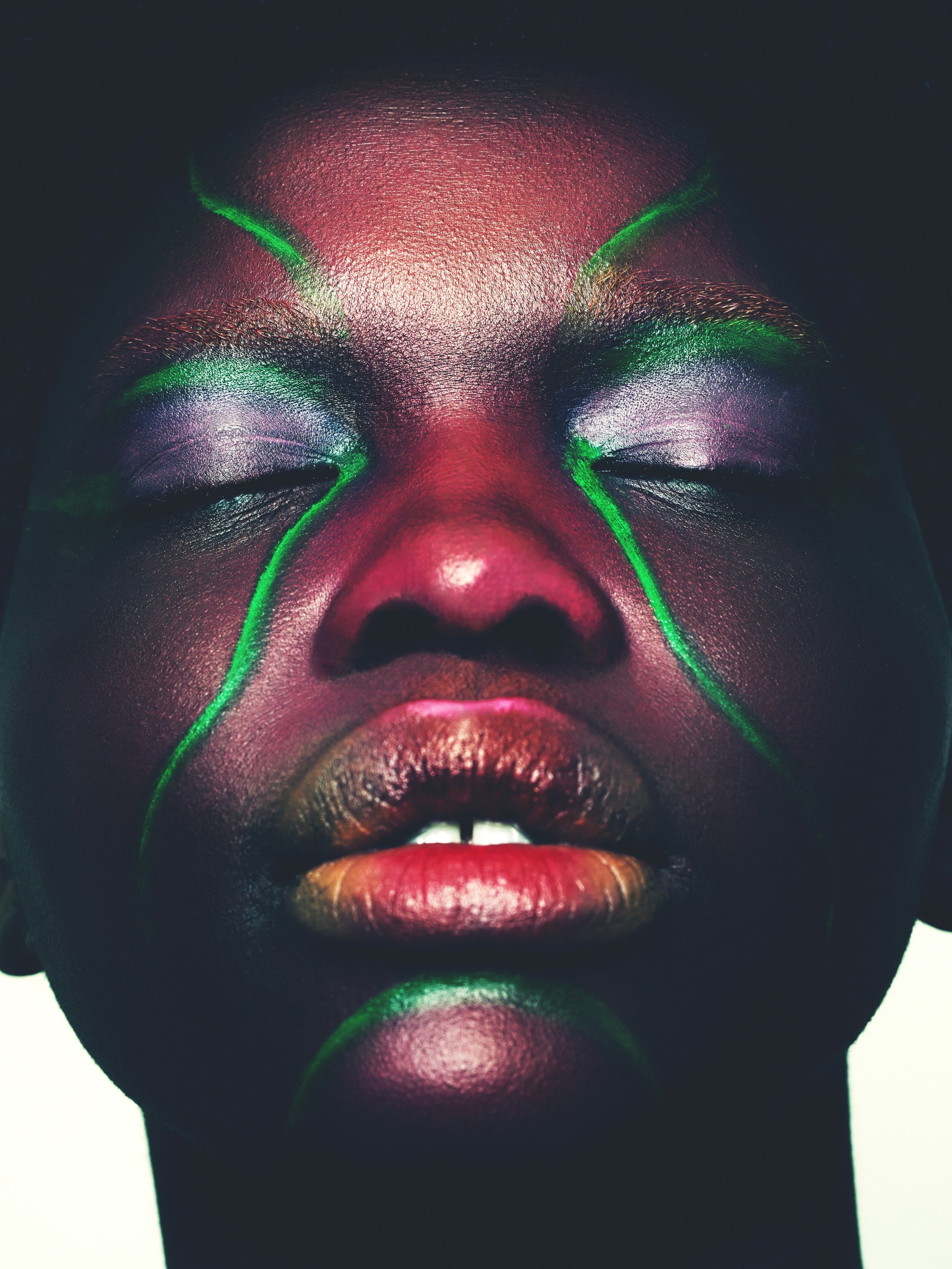 Close-up portrait of a person with dark skin and closed eyes, enhanced with green facial paint and iridescent eye makeup for a beauty shot.