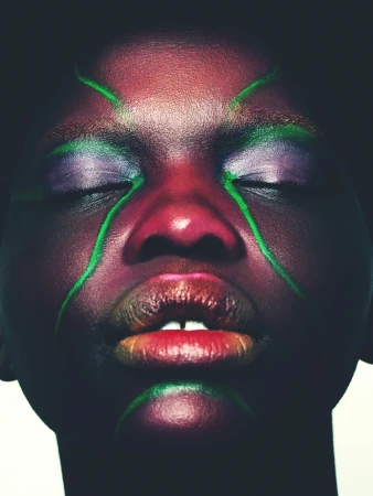 Close-up portrait of a person with dark skin and closed eyes, enhanced with green facial paint and iridescent eye makeup for a beauty shot.