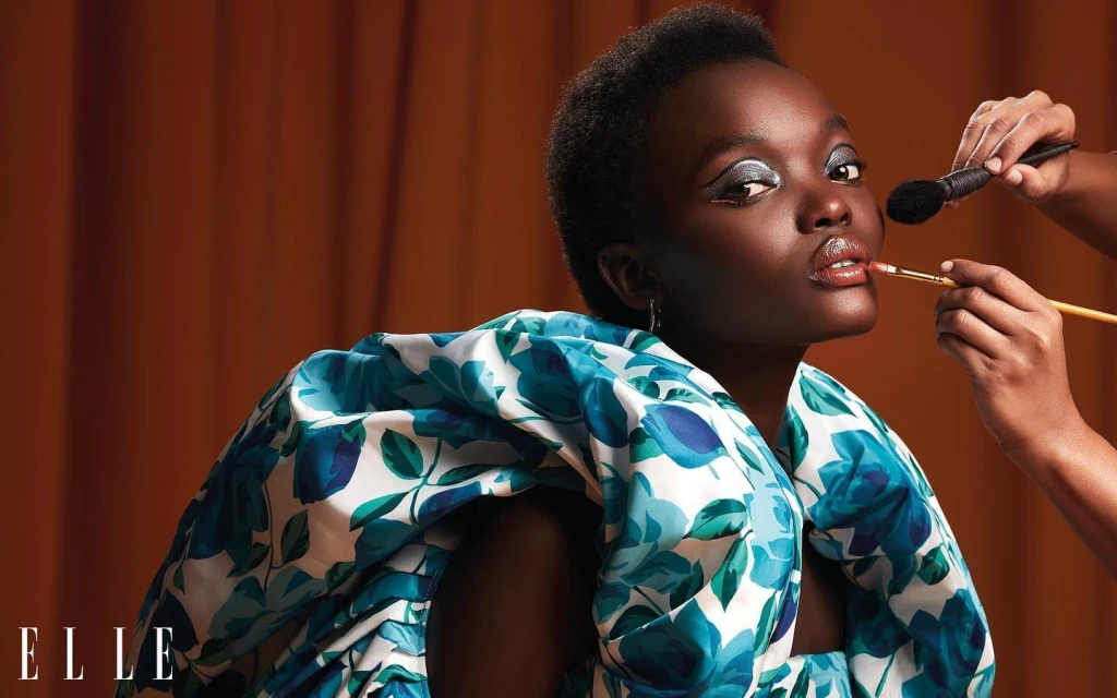 Glamorous beauty shot of a young Black woman having her makeup applied by a makeup artist in a studio setting with brown backdrop.