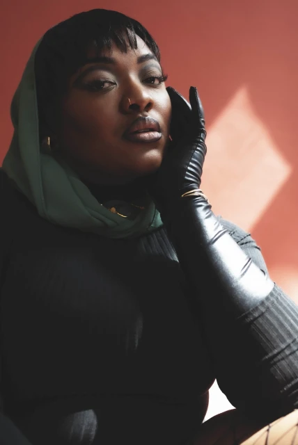Portrait of an adult Black woman with bangs, wearing a green headscarf, black top, and black gloves, against a peach backdrop.