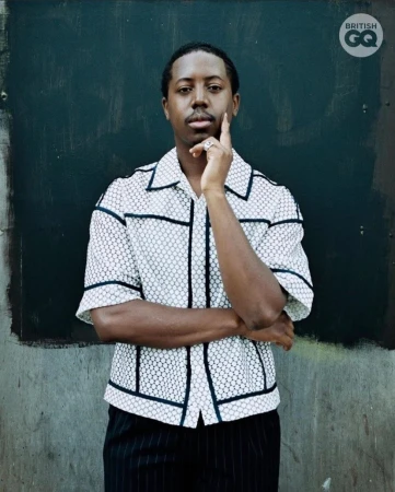 Portrait of a black man wearing a white patterned short-sleeve shirt with dark trim against a dark textured wall