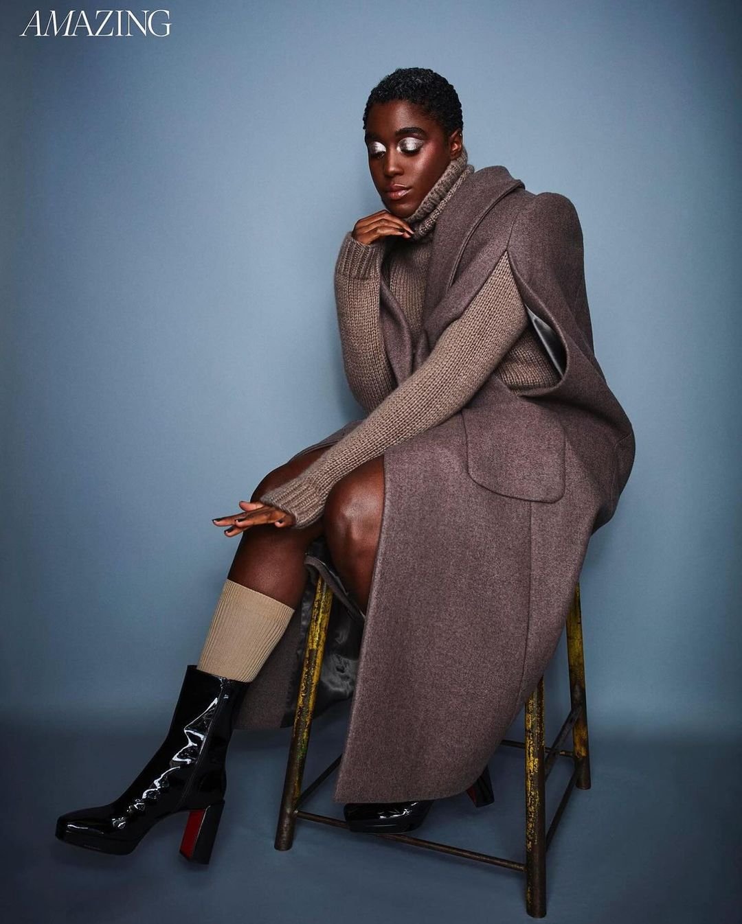 A single Black woman sits on a stool wearing a brown turtleneck sweater, long wool coat, beige socks, and shiny black boots in a photo studio.