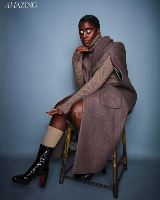 A single Black woman sits on a stool wearing a brown turtleneck sweater, long wool coat, beige socks, and shiny black boots in a photo studio.