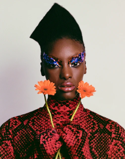 Portrait of Black woman with creative makeup, wearing a red snakeskin print top, holding orange flowers in a photo studio setting.