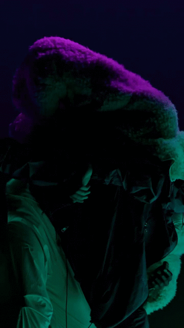 Two individuals pose in vibrant green and purple lighting, showcasing a unique fur coat in an indoor setting.