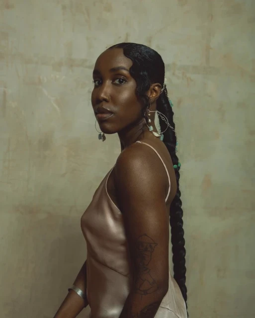 Portrait of an adult woman with braided hair and a light pink dress posing in front of a textured mottled wall.