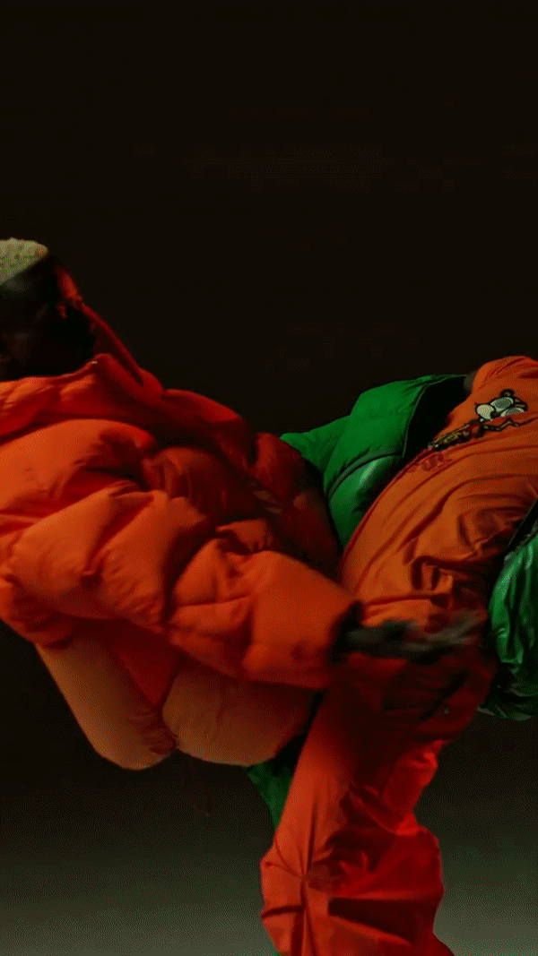 Two people wear brightly colored green and orange puffer tracksuits in a full shot on a plain dark backdrop.