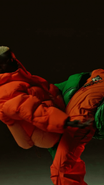 Two people wear brightly colored green and orange puffer tracksuits in a full shot on a plain dark backdrop.